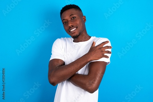 Charming pleased young handsome man wearing white T-shirt over blue background embraces own body, pleasantly feels comfortable poses. Tenderness and self esteem concept