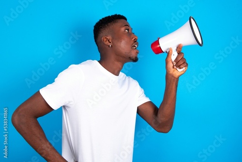 Funny young handsome man wearing white T-shirt over blue background People sincere emotions lifestyle concept. Mock up copy space. Screaming in megaphone.