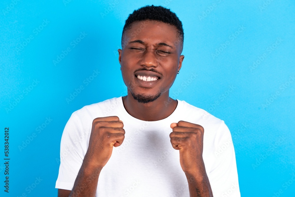 young handsome man wearing white T-shirt over blue background grins joyfully, imagines something pleasant, copy space. Pleasant emotions concept. young handsome man wearing white T-shirt over blue background grins joyfully, imagines something pleasant, copy space. Pleasant emotions concept.