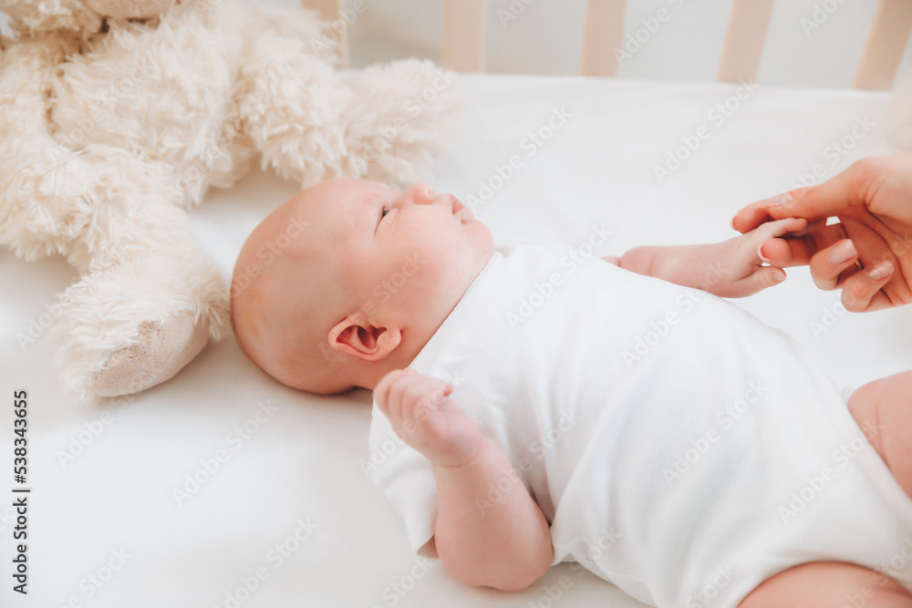 A charming smiling blue-eyed 2-month-old baby in a white bodysuit lies in a crib next to a teddy bear. View from above. a newborn. A charming smiling blue-eyed 2-month-old baby in a white bodysuit lies in a crib next to a teddy bear. View from above. a newborn.