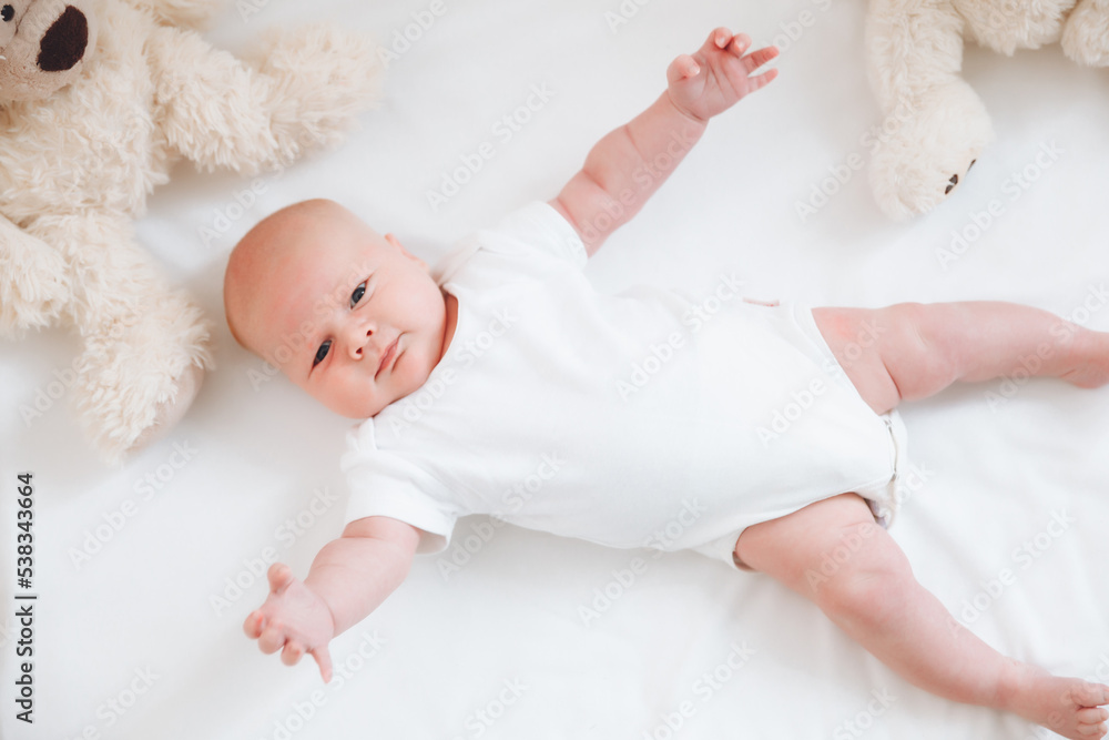 A charming smiling blue-eyed 2-month-old baby in a white bodysuit lies in a crib next to a teddy bear. View from above. a newborn.
