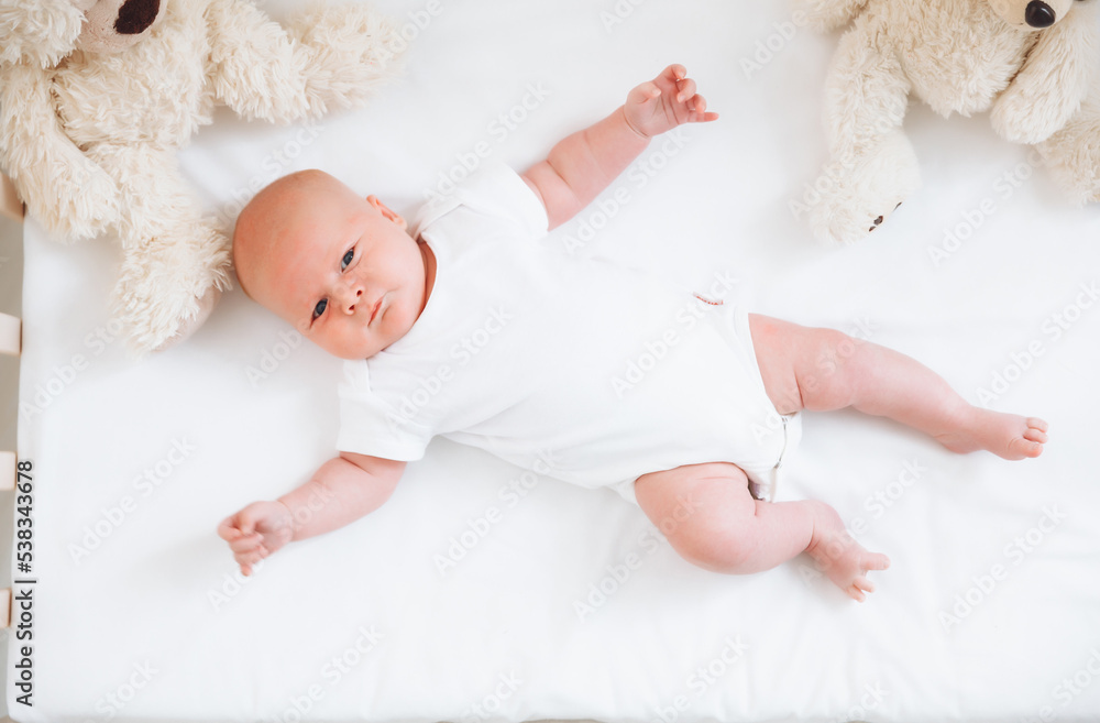 A charming smiling blue-eyed 2-month-old baby in a white bodysuit lies in a crib next to a teddy bear. View from above. a newborn. A charming smiling blue-eyed 2-month-old baby in a white bodysuit lies in a crib next to a teddy bear. View from above. a newborn.