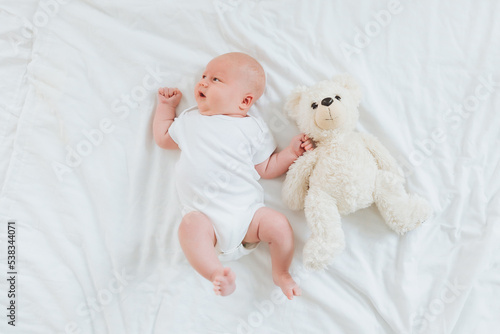 a 2-month-old baby in a white bodysuit is lying at home on a white bed next to a teddy bear. view from above. newborn