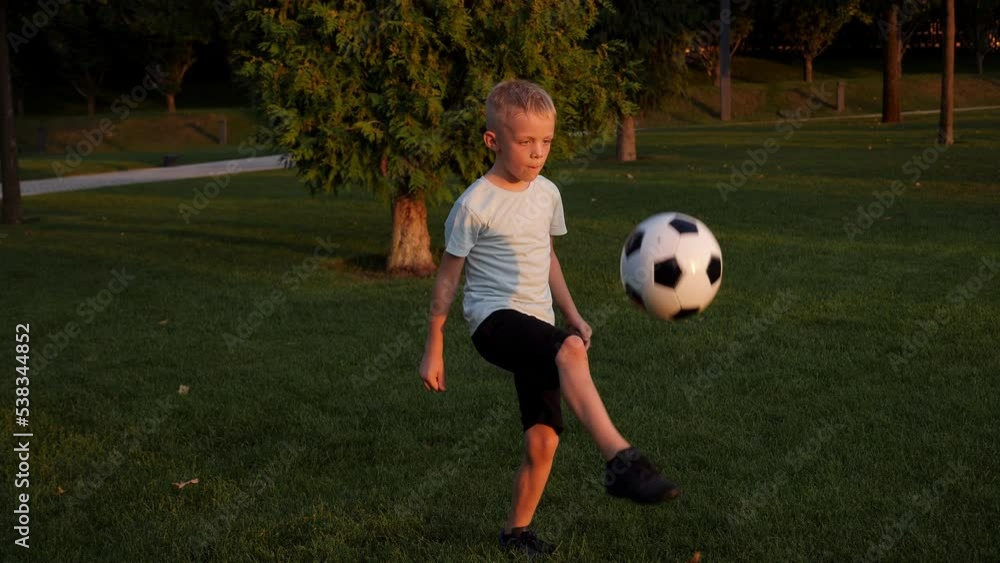 A little athletic boy trains and stuffs a ball on the green grass in the city park in the setting sun.