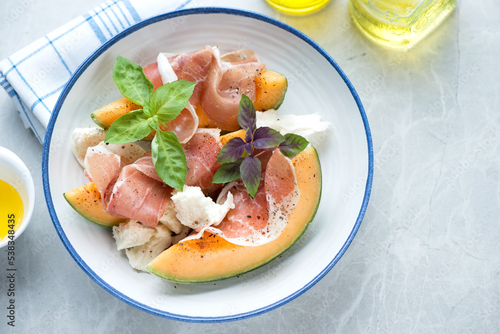Plate with cantaloupe slices, prosciutto, mozzarella and basil, high angle view on a light-grey granite background, horizontal shot Plate with cantaloupe slices, prosciutto, mozzarella and basil, high angle view on a light-grey granite background, horizontal shot