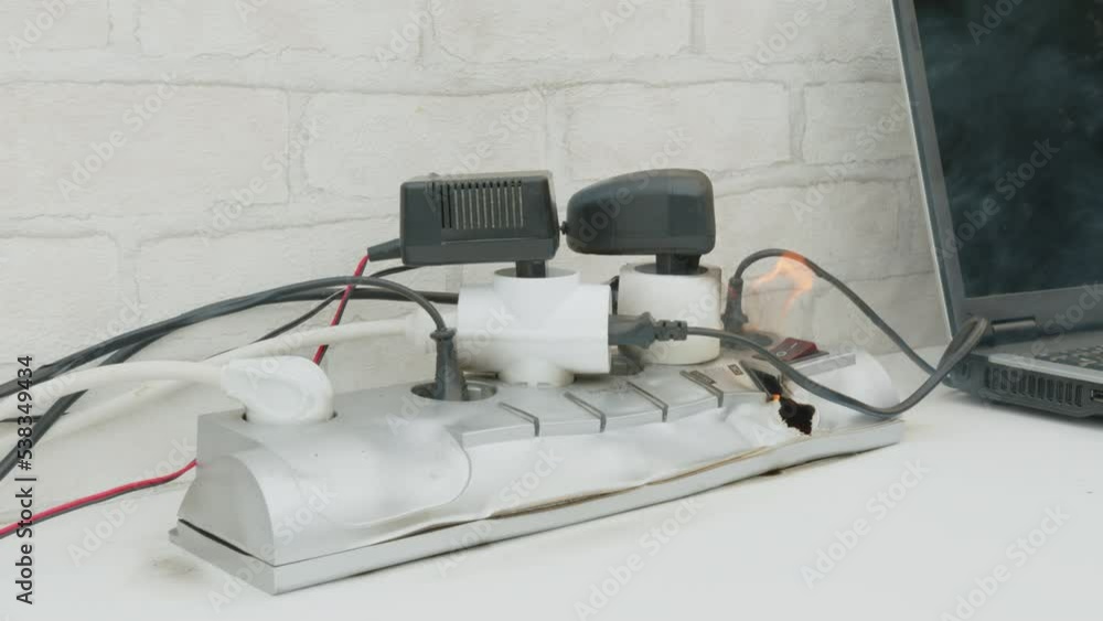 Overloaded power cube on fire on table indoors close up, voltage overload. Lot of electric plugs in sockets of power strip.
