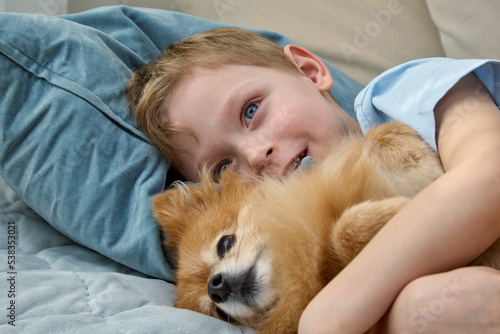 Close-up of a smiling blond child gently hugging a cute red dog. A happy boy with his German Spitz is lying on a bed with pillows. Friendship with a charming pet. © Aleksandr