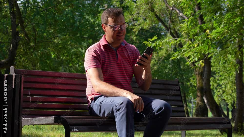 Portrait of mature caucasian man in glasses sitting on park bench holding mobile phone and waving hello. Handsome guy in pink shirt talking on video call in public place. Portrait of mature caucasian man in glasses sitting on park bench holding mobile phone and waving hello. Handsome guy in pink shirt talking on video call in public place.