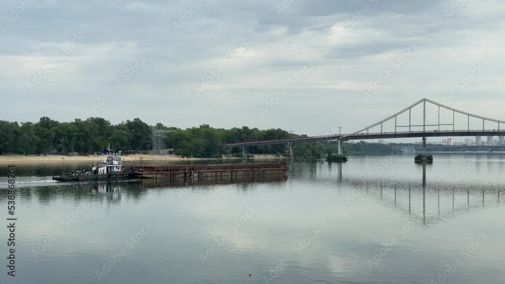 Industrial tugboat towing a long barge with sand, sailing along the fairway of the river at sunset. On the horizon, two bridges across the Dnieper River in Kyiv Industrial tugboat towing a long barge with sand, sailing along the fairway of the river at sunset. On the horizon, two bridges across the Dnieper River in Kyiv