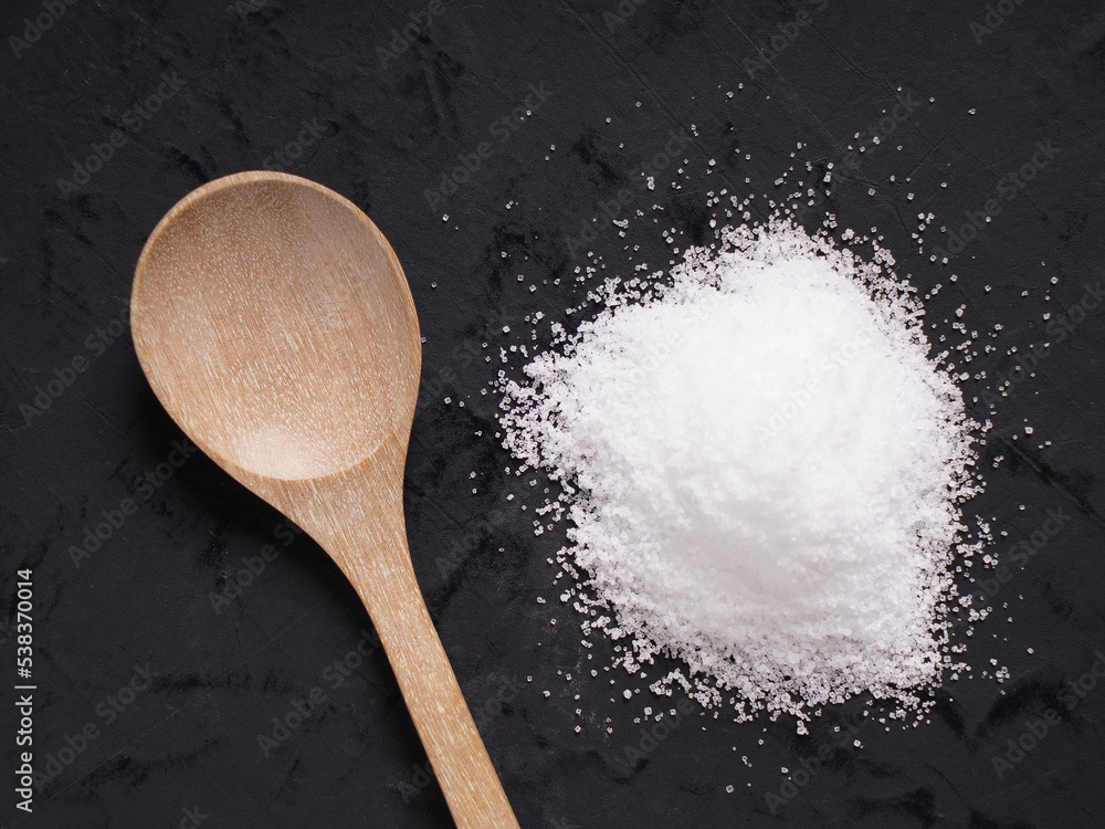White table salt is placed on a black stone background with an old wooden spoon.