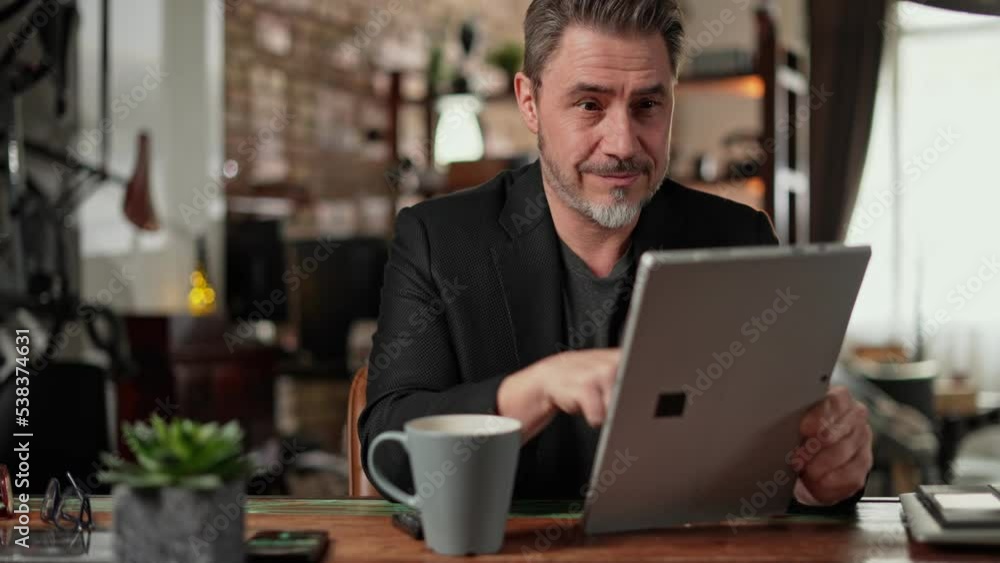 Older man sitting at desk in dark cosy room working on tablet computer in home office. Mature age, middle age, mid adult casual man in 50s, confident happy smiling. Older man sitting at desk in dark cosy room working on tablet computer in home office. Mature age, middle age, mid adult casual man in 50s, confident happy smiling.