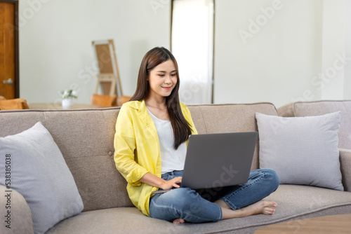 Young woman is sitting with squat on comfortable couch and putting laptop on the legs to typing business