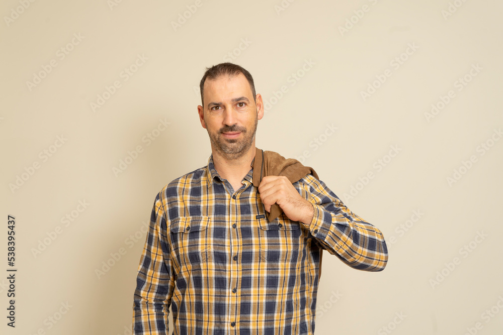 Latin handsome bearded man posing in a checkered shirt with a brown jacket on his shoulder, isolated over beige background. Latin handsome bearded man posing in a checkered shirt with a brown jacket on his shoulder, isolated over beige background.