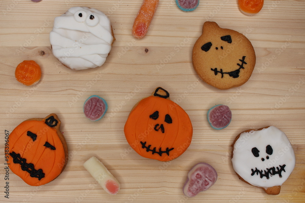 Spooky Halloween cookies on a light wood table. White and orange fondant and icing. There are sweets and candies decorating the background. Wallpaper of a party, celebration and festivity. Spooky Halloween cookies on a light wood table. White and orange fondant and icing. There are sweets and candies decorating the background. Wallpaper of a party, celebration and festivity.