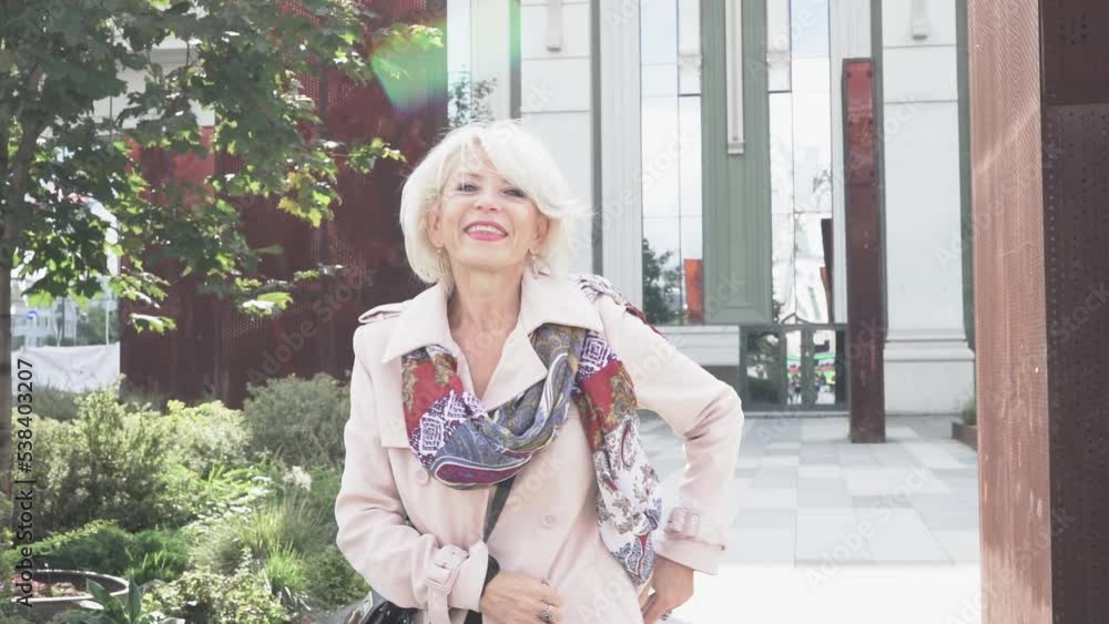 A happy mature woman with silver hair in slow motion, strolling along a narrow street against the background of a mirrored building. High quality FullHD footage A happy mature woman with silver hair in slow motion, strolling along a narrow street against the background of a mirrored building. High quality FullHD footage