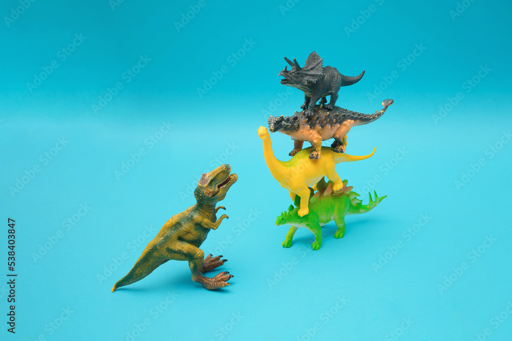 Fototapeta premium Four dinosaurs stand on each other on a blue background. Ankylosaurus, Brachiosaurus, Stegosaurus and Triceratops against T-Rex. Blue background. Minimal design and concept..Plastic toys.