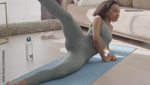Slowmo of attractive young Biracial woman doing side leg raise exercise on mat in sunlit living room photo