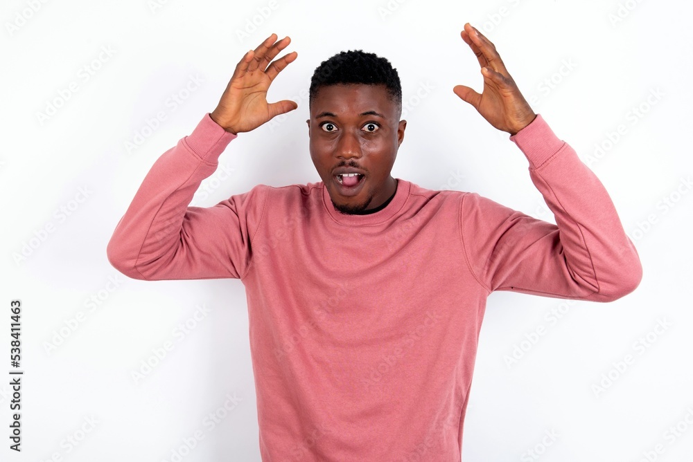 young handsome man wearing pink sweater over white background looks with excitement at camera, keeps hands raised over head, notices something unexpected, reacts on sudden news.