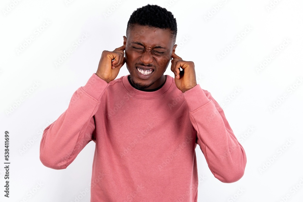 Stop making this annoying sound! Unhappy stressed out young handsome man wearing pink sweater over white background making worry face, plugging ears with fingers, irritated with loud noise. Stop making this annoying sound! Unhappy stressed out young handsome man wearing pink sweater over white background making worry face, plugging ears with fingers, irritated with loud noise.