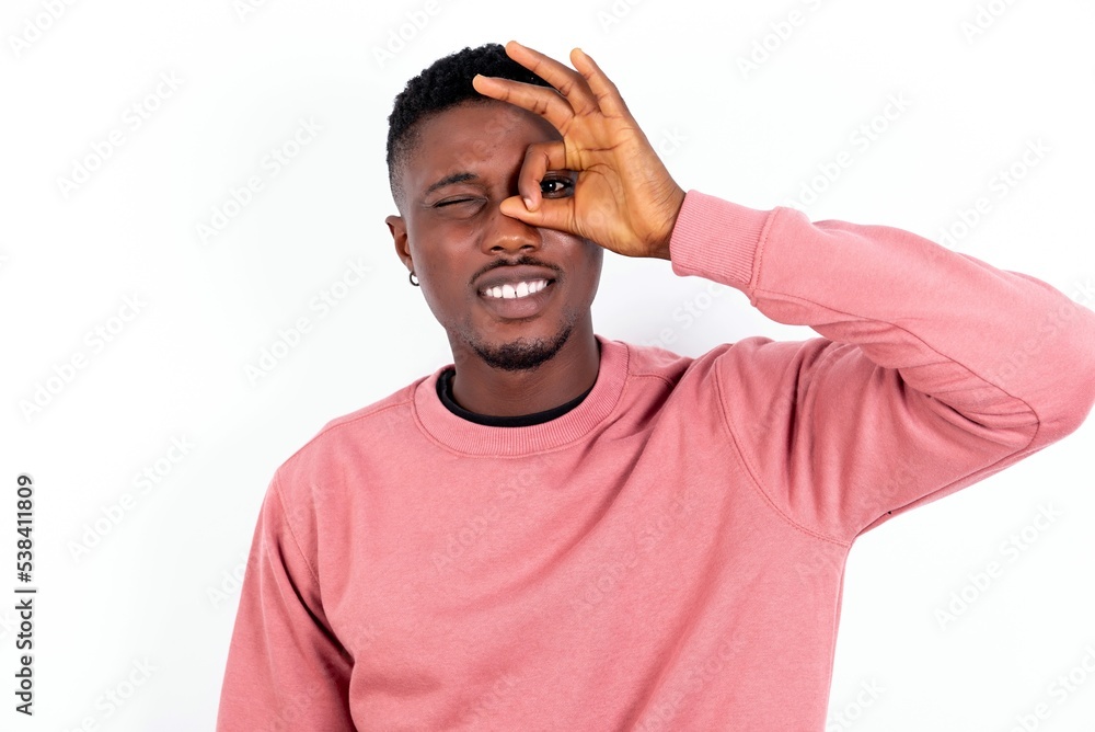 young handsome man wearing pink sweater over white background with happy face smiling doing ok sign with hand on eye looking through finger. young handsome man wearing pink sweater over white background with happy face smiling doing ok sign with hand on eye looking through finger.