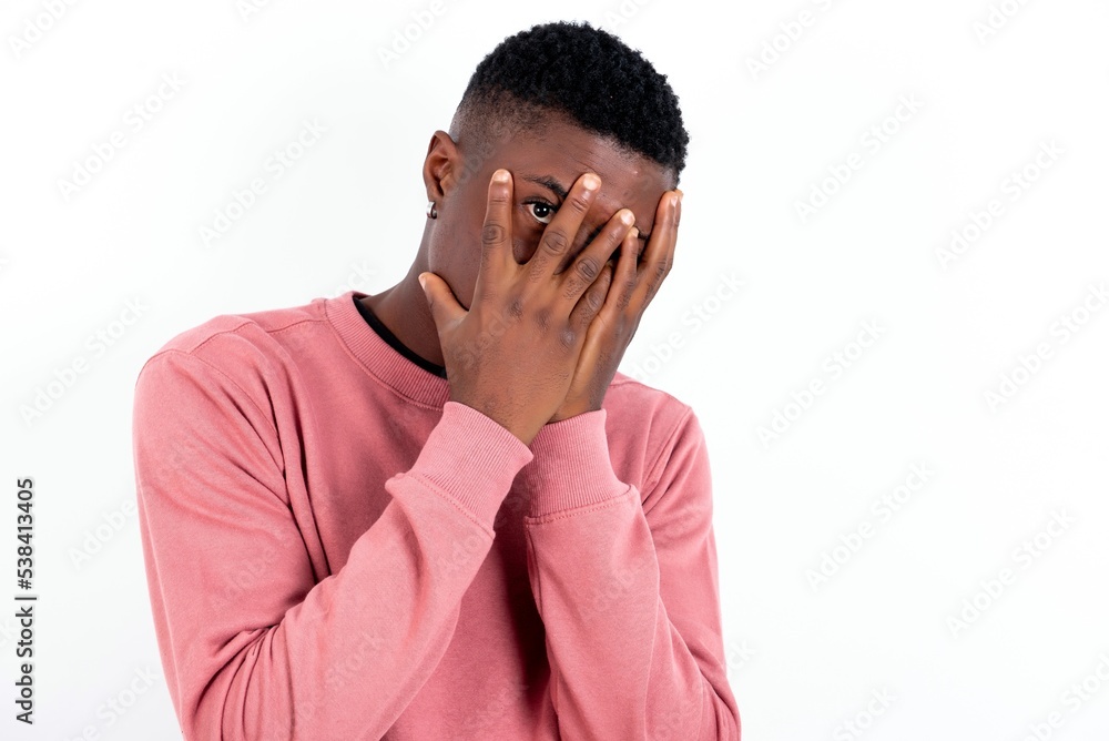young handsome man wearing pink sweater over white background covering face with hands and peering out with one eye between fingers. Scared from something or someone. young handsome man wearing pink sweater over white background covering face with hands and peering out with one eye between fingers. Scared from something or someone.