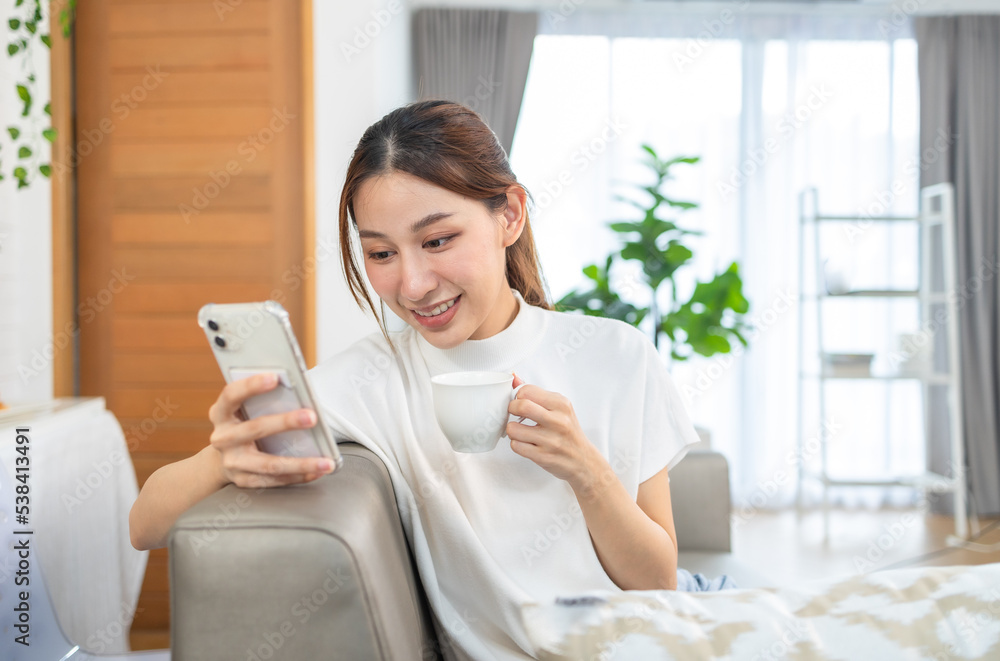 Happy and cheerful woman is using a phone sitting on a sofa.Asian young woman looking mobile phone for shopping online at home. Happy and cheerful woman is using a phone sitting on a sofa.Asian young woman looking mobile phone for shopping online at home.