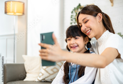 Young asian mother and her daughter are making selfie using a phone, hugging and smiling while sitting on the sofa at home.
