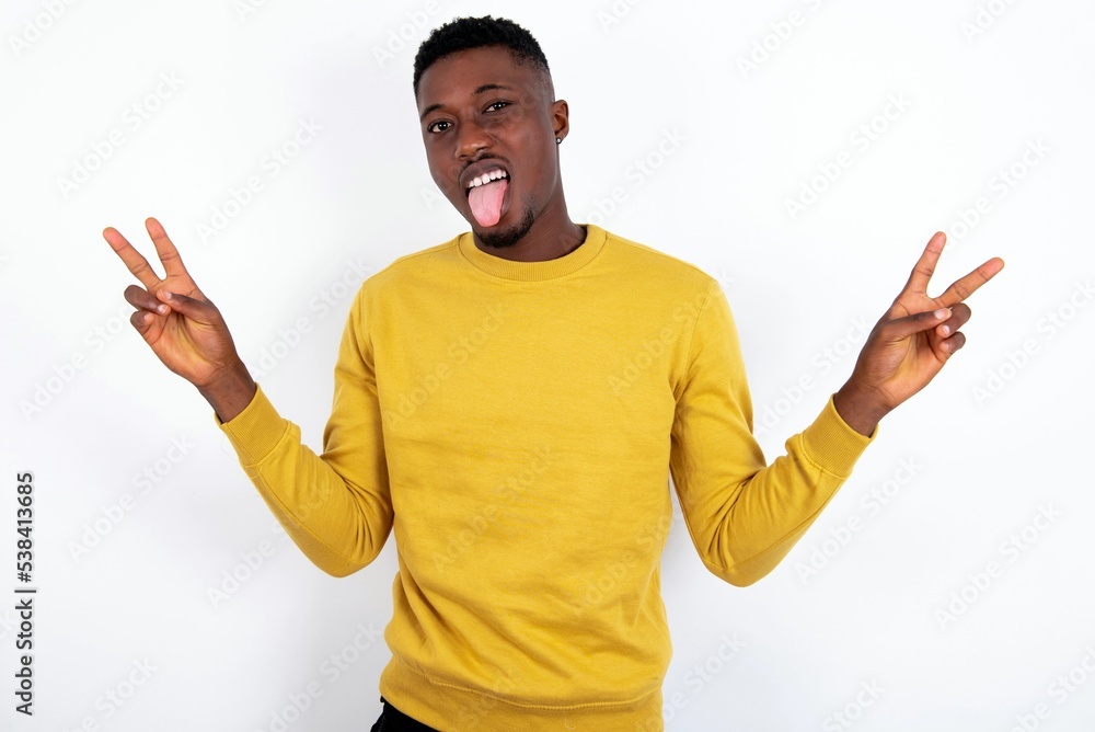young handsome man wearing yellow sweater over white background with optimistic smile, showing peace or victory gesture with both hands, looking friendly. V sign. young handsome man wearing yellow sweater over white background with optimistic smile, showing peace or victory gesture with both hands, looking friendly. V sign.