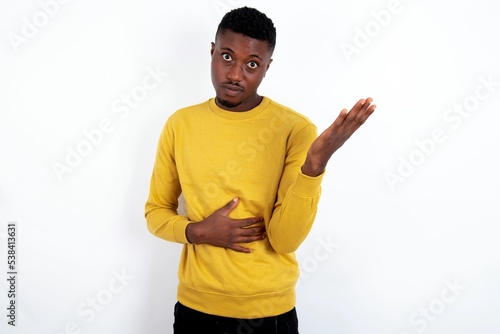 Studio shot of frustrated young handsome man wearing yellow sweater over white background gesturing with raised palm, frowning, being displeased and confused with dumb question.