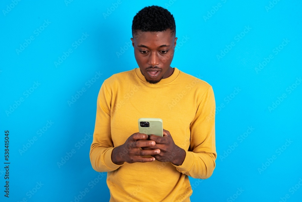 young handsome man wearing yellow T-shirt over blue background using mobile phone chatting free time . young handsome man wearing yellow T-shirt over blue background using mobile phone chatting free time .