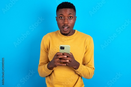 Excited young handsome man wearing yellow T-shirt over blue background holding smartphone and looking amazed to the camera after receiving good news.