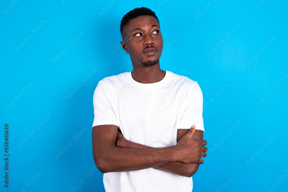 Fototapeta premium Pleased young handsome man wearing white T-shirt over blue background keeps hands crossed over chest looks happily aside