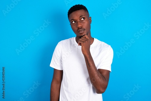 Thoughtful young handsome man wearing white T-shirt over blue background holds chin and looks away pensively makes up great plan