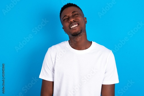 Positive young handsome man wearing white T-shirt over blue background with overjoyed expression closes eyes and laughs shows white perfect teeth