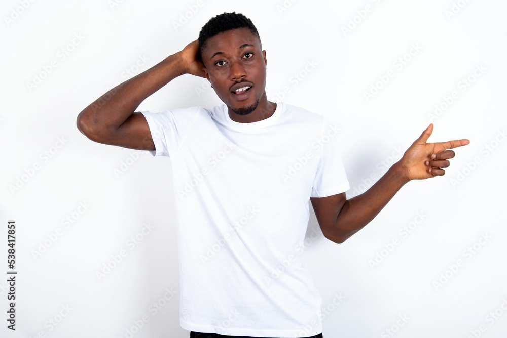 Surprised young handsome man wearing white T-shirt over white background pointing at empty space holding hand on head Surprised young handsome man wearing white T-shirt over white background pointing at empty space holding hand on head
