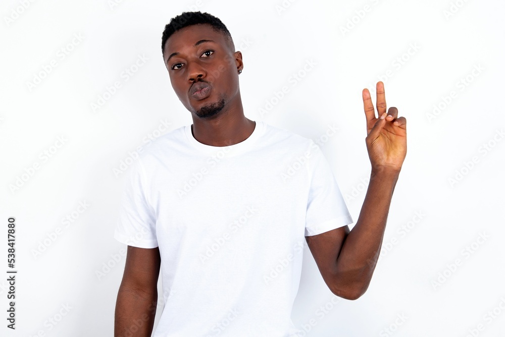 young handsome man wearing white T-shirt over white background makes peace gesture keeps lips folded shows v sign. Body language concept young handsome man wearing white T-shirt over white background makes peace gesture keeps lips folded shows v sign. Body language concept