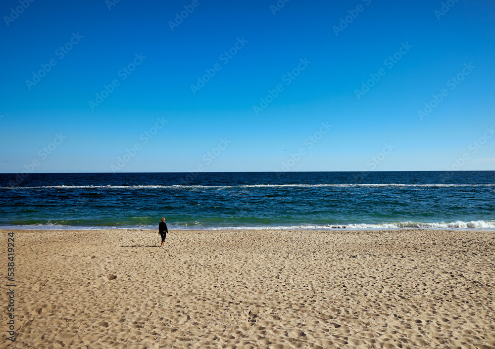 Huge beach, blue sea and sky and a lonely young woman walking along the beach towards the sea