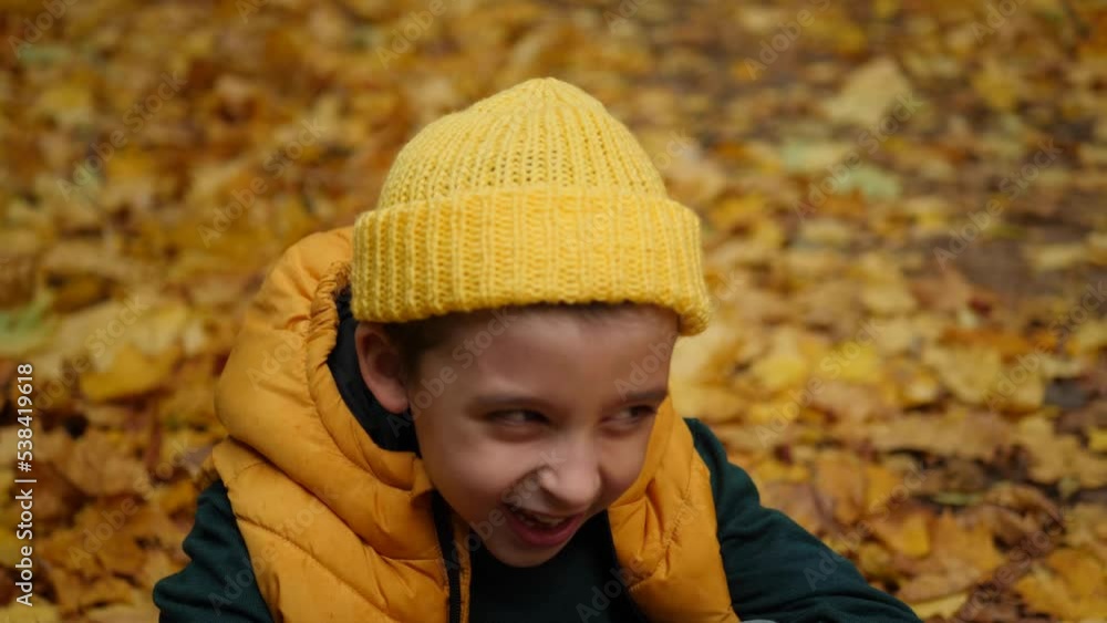 portrait of a funny boy child without 2 teeth sitting and laughter in autumn in maple leaves nature. he is wearing a yellow vest, a knitted hat and a green sweater portrait of a funny boy child without 2 teeth sitting and laughter in autumn in maple leaves nature. he is wearing a yellow vest, a knitted hat and a green sweater