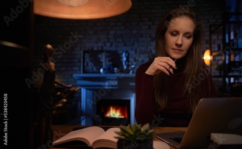 Home office - white woman working from home with laptop computer. Sitting at desk in dark living room in front of fireplace in Winter.