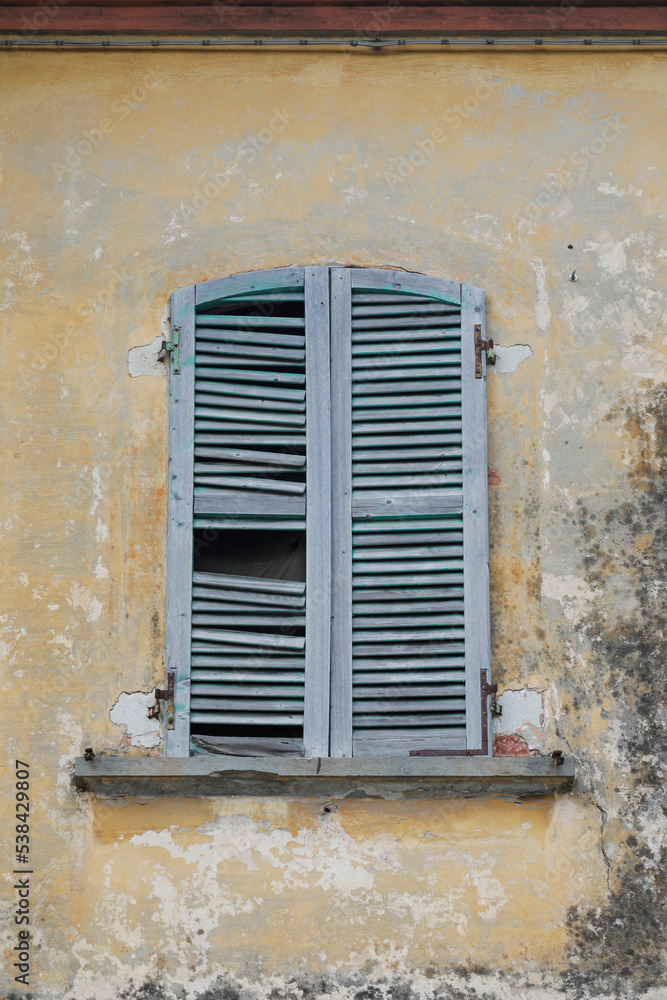 Damaged old window with shutters on a very ancient house wall, wall dirty and frame cracked, vertical format
