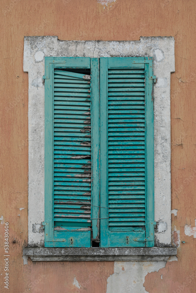 Window with closed green damaged wooden shutters,
orange and grey house wall with rough surface, no person, vertical format