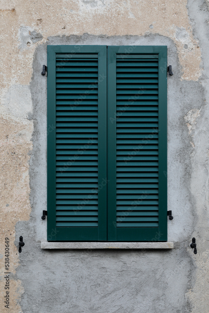Cracked cement wall with window and closed wooden shutter in green, vertical format