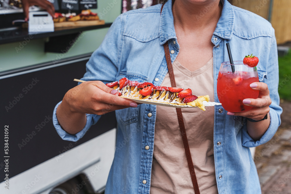 Woman enjoys delicious pancakes with strawberries and strawberry daiquiri at a food festival. Crepes with fresh strawberries and whipped cream, street food. Food trucks, food festival.