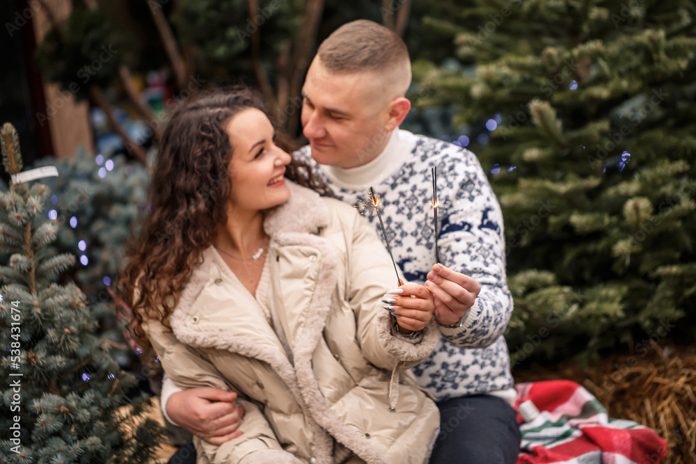 Fototapeta premium A beautiful young couple sitting near Christmas trees in white clothes walking near garlands. Happy man and woman, romance, Christmas celebration, fun, love. White sweater, new year, street, outside