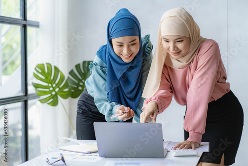 Two beautiful Asian Muslim women wearing headscarves working in the office with laptops with financial graphs. Muslim businesswoman in traditional dress working and talking in office meeting