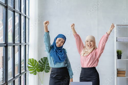 Two beautiful Asian Muslim business women wearing headscarves celebrate their achievements by clapping each other's hands and raising their hands.