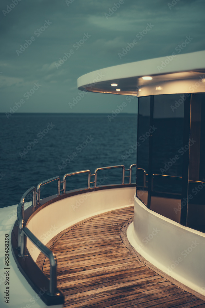 Fototapeta premium A vertical low-key view of a diving safari yacht's middle deck with a chrome fence, lit wooden floor, illuminated ceiling, and windows reflecting the dark ocean waterscape and part of the evening sky
