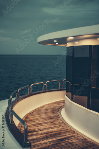 A vertical low-key view of a diving safari yacht's middle deck with a chrome fence, lit wooden floor, illuminated ceiling, and windows reflecting the dark ocean waterscape and part of the evening sky © skyNext
