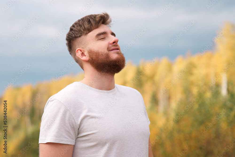 Young happy calm relaxed bearded man enjoying good weather in the forest, park, breathing deep inhale deeply fresh air and smiling. Handsome guy with beard on natural background with his eyes closed