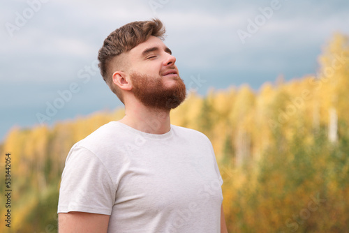 Young happy calm relaxed bearded man enjoying good weather in the forest, park, breathing deep inhale deeply fresh air and smiling. Handsome guy with beard on natural background with his eyes closed